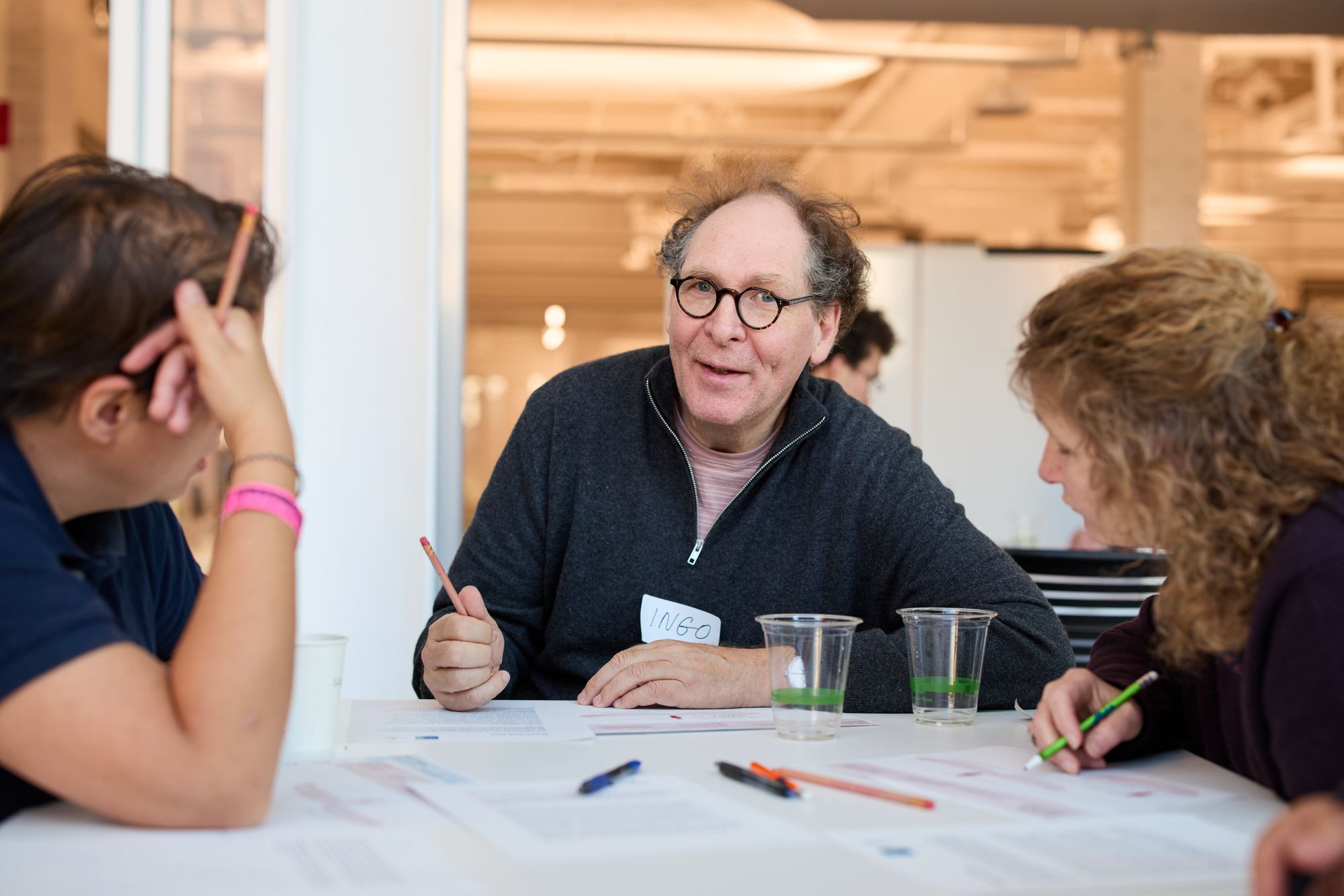 A group discusses, smiling, while they look at a spread of papers and pens.